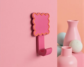 May include: A pink wall hook with a scalloped top and a yellow outline. The hook is mounted on a pink wall. Decorative pink vases and spheres are in the background.