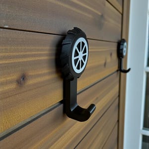 Peut inclure: Crochets muraux en métal noir avec un design de roue. Les crochets sont montés sur un mur en bois. Le design de la roue est noir avec un motif de rayons blancs. Solution de rangement décorative et fonctionnelle.