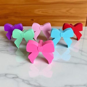 May include: A selection of colourful bow-shaped bottle stoppers in shades of purple, green, pink, blue, and red. The stoppers are arranged on a white marble surface, with a wooden background.