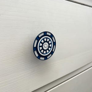 Könnte beinhalten: Runder Schrankknauf mit blauem und weißem geometrischem Design. Der Knauf ist an einer weißen Holzschublade befestigt. Der Knauf hat eine kreisförmige Form mit einer Reihe konzentrischer Ringe und einem zentralen Muster. Durchmesser: ca. 5 cm.