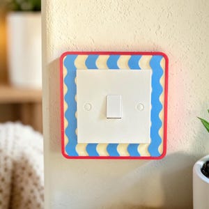 May include: A decorative light switch cover with a red border, blue wave pattern, and a white center. The switch is white and rectangular. The cover is mounted on a cream-colored wall.