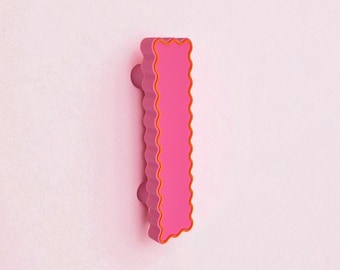 May include: A pink cabinet pull with a wavy edge and orange trim. The pull has a cylindrical knob on each end. The hardware is mounted on a light pink surface. This decorative pull is a unique accent for cabinets and drawers.