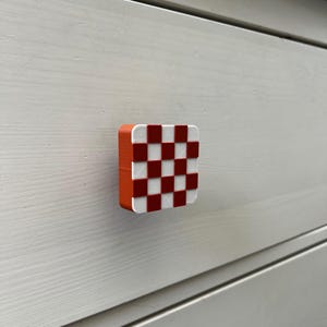Op de afbeelding: Vierkante kastknop met een rood-wit dambordpatroon aan de voorkant. De knop heeft een oranje rand en is gemonteerd op een wit houten oppervlak. Deze decoratieve knop voegt kleur toe.