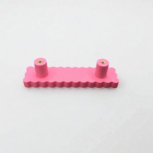 Puede incluir: Un tirador de armario rosa con un dise&ntilde;o de borde festoneado. El tirador tiene dos pomos cil&iacute;ndricos con inserciones met&aacute;licas doradas. El herraje est&aacute; dise&ntilde;ado para muebles y a&ntilde;ade un toque decorativo.