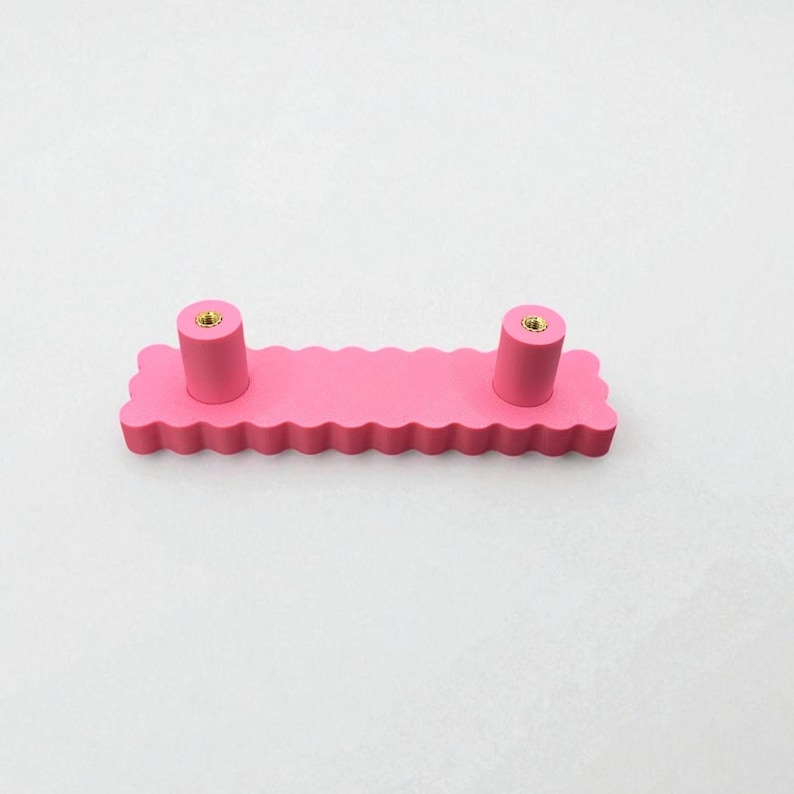 Puede incluir: Un tirador de armario rosa con un dise&ntilde;o de borde festoneado. El tirador tiene dos pomos cil&iacute;ndricos con inserciones met&aacute;licas doradas. El herraje est&aacute; dise&ntilde;ado para muebles y a&ntilde;ade un toque decorativo.