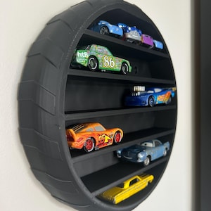 May include: A black, circular tyre-shaped shelf unit displaying toy cars. The shelf has multiple levels, each holding a different toy car. The cars are various colours, including blue, green, orange, and yellow. The shelf is mounted on a white wall.