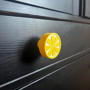 Könnte beinhalten: Gelber Schrankknauf in Form einer Zitronenscheibe mit weißen Akzenten und einem gelben Zentrum. Der Knauf ist an einem schwarzen Holzschrank befestigt und verleiht den Möbeln einen Farbtupfer und ein Zitrus-Thema.