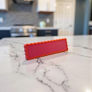 May include: A rectangular, pink label with an orange wavy border. The label is made of a solid material and is displayed on a white marble surface. The label is a vibrant accent piece.