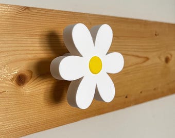 May include: A white daisy-shaped decorative knob with a yellow center is mounted on a light brown wooden plank. The knob is three-dimensional and has a simple, clean design. The wood grain is visible, adding a natural element to the piece.