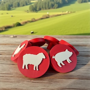 Op de afbeelding: Een verzameling rode, ronde houten flessenstoppers met witte dierensilhouetten. De stoppers hebben een schaap-, varken- en kippenontwerp. De achtergrond toont een houten oppervlak en een groen veld.