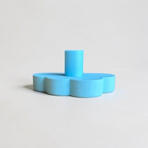 May include: A bright sky blue candle holder shaped like a stylized flower with rounded petals. The base is flat and wide, supporting a central cylindrical opening designed to hold a single taper candle. The surface has a smooth, matte finish.