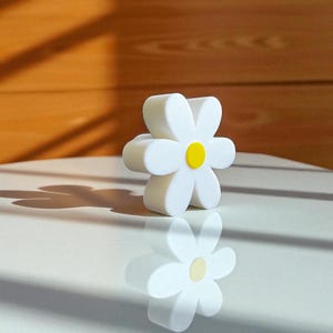 May include: A white daisy-shaped object with a yellow centre. The object is on a white surface, with its reflection visible. The background is a light brown wooden wall. The daisy has six petals.