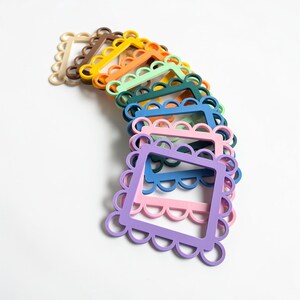May include: A collection of colorful, scalloped-edge picture frames in various shades, including beige, brown, yellow, orange, green, blue, pink, and purple. The frames are stacked and arranged on a white surface.
