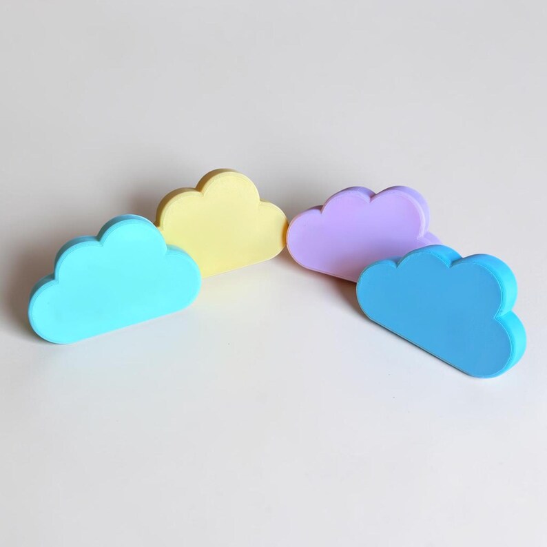 May include: Four cloud-shaped objects in pastel colors, including one light blue, one yellow, one lavender, and one medium blue cloud. Each piece has a smooth, matte finish and a rounded, scalloped edge design, arranged in a semi-circular formation.