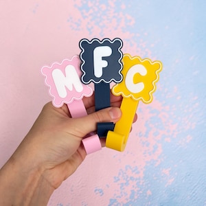 Könnte beinhalten: Drei farbenfrohe Buchstabenclips werden in einer Hand gehalten. Die Buchstaben sind M, F und C. Das M ist rosa mit einem weißen Buchstaben, das F ist marineblau mit einem weißen Buchstaben und das C ist gelb mit einem weißen Buchstaben. Die Clips haben einen gewellten Rand.