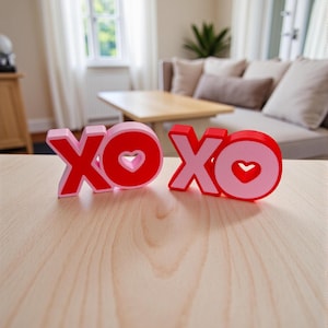 Puede incluir: Dos letreros decorativos de madera XOXO en rojo y rosa, con recortes en forma de corazón en las O. Las letras están colocadas sobre una superficie de madera de color claro, con un fondo borroso de sala de estar.