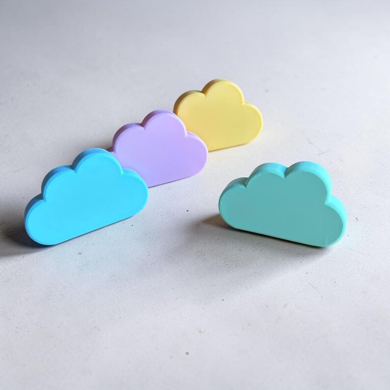 May include: Four cloud-shaped objects in pastel colors, including bright blue, light purple, soft yellow, and mint green. Each piece has a smooth, matte finish and a rounded, scalloped top edge, standing upright on a flat, neutral-toned surface.