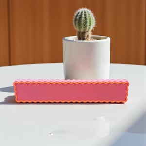 Könnte beinhalten: Ein rechteckiges rosa Objekt mit einem wellenförmigen orangefarbenen Rand. Ein kleiner Kaktus in einem weißen Topf befindet sich im Hintergrund. Die Objekte befinden sich auf einer weißen Oberfläche mit braunem Hintergrund.