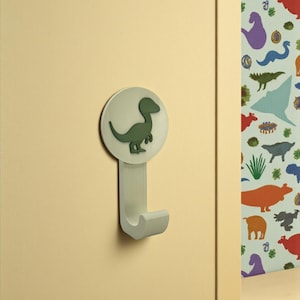 Può includere: Un gancio da parete verde chiaro con un design a dinosauro. Il gancio presenta una base circolare con una silhouette di dinosauro verde e un gancio curvo. È montato su una parete giallo chiaro, con carta da parati a tema dinosauri sullo sfondo.