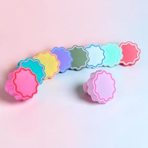 Könnte beinhalten: Eine Sammlung von neun runden Knöpfen mit gewelltem Rand, die jeweils eine eigene Volltonfarbe aufweisen, darunter Pink, Türkis, Gelb, Lila, Olivgrün, Hellblau, Mintgrün, Koralle und Lavendel, angeordnet in einer geschwungenen Linie.