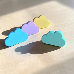 May include: Four cloud-shaped objects in pastel colors, including light blue, lavender, pale yellow, and mint green. Each piece has a smooth, solid finish and a rounded, multi-lobed top edge, arranged in a staggered formation on a flat surface.