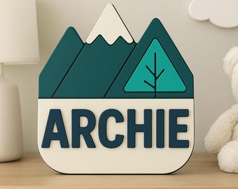 May include: A personalized name sign with a mountain and tree design. The sign features the name "ARCHIE" in navy blue letters on a cream-colored base. The mountain and tree are teal and white.