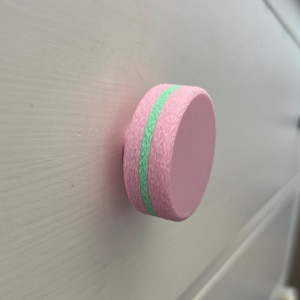 May include: A pink cabinet knob with a textured surface and a light green stripe. The round knob is attached to a white cabinet door. This decorative knob adds a pop of colour to any furniture piece.