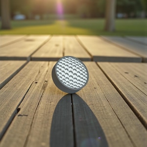 May include: A grey, circular object with a white, patterned top, resting on a wooden surface. The pattern consists of numerous small, curved shapes. The object casts a shadow on the wooden planks, with a blurred background of trees and sunlight.