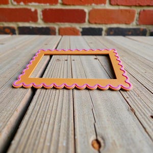 May include: An orange, scalloped-edge picture frame with a pink border. The frame is rectangular and sits on a weathered wooden surface, with a brick wall in the background. The frame is empty.