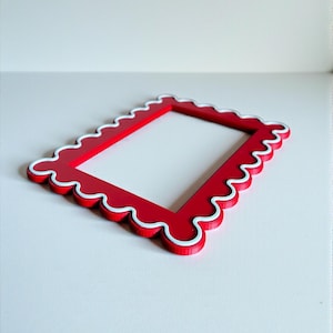 Puede incluir: Un marco de fotos rectangular rojo con borde festoneado y ribete blanco. El marco es de madera y está diseñado para contener una fotografía o una obra de arte. El marco es de color rojo vibrante.