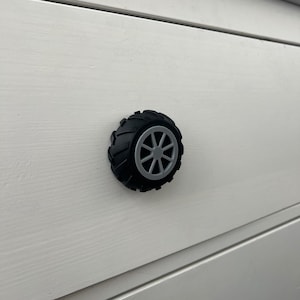 May include: A black and grey tyre-shaped cabinet knob. The knob features a detailed tyre tread design with a grey wheel centre in a starburst pattern. The knob is mounted on a white wooden cabinet door.