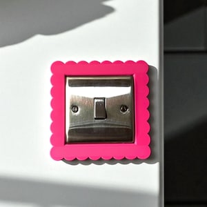 Può includere: Un interruttore in acciaio inossidabile con un interruttore a levetta, incorniciato da una cornice rosa brillante con bordo smerlato. L'interruttore è montato su una parete bianca, con ombre. Un elemento decorativo e funzionale per la casa.