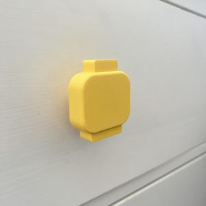 May include: A bright yellow cabinet knob shaped like a toy brick head. The knob is mounted on a white surface. The knob has a rectangular base and a rounded top.