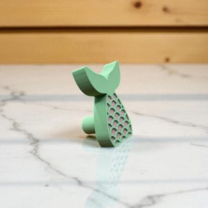 May include: A mint green mermaid tail-shaped cabinet knob with a pink scale pattern. The knob has a rounded base for easy installation and a decorative tail design. This unique hardware adds a whimsical touch to furniture.