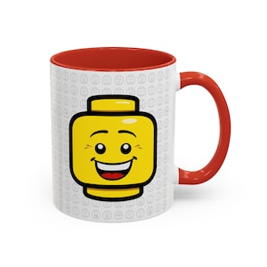 Könnte beinhalten: Eine weiße Keramiktasse mit rotem Rand und Henkel. Die Tasse zeigt einen gelben Lego-Kopf mit einem lächelnden Gesicht auf weißem Hintergrund mit einem Muster aus kleinen Lego-Köpfen.