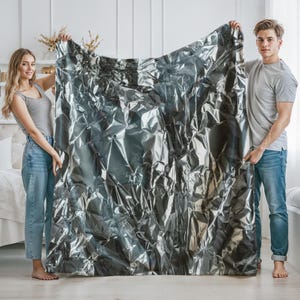 May include: Two people holding up a large sheet of crumpled silver foil. The foil is textured and has a metallic sheen.