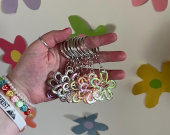 May include: A hand holds a collection of flower-shaped keychains. Each keychain features a clear, iridescent flower charm attached to a silver metal ring. The background includes colorful paper flower cutouts.
