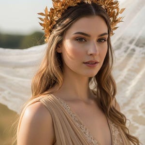 May include: A woman wearing a gold leaf crown and a beige dress. The crown is made of gold-colored leaves and sits atop her long, wavy brown hair. A sheer, white fabric flows in the background. The dress has a V-neckline with embroidered details.
