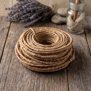 Könnte beinhalten: Ein aufgerolltes Stück naturfarbenes braunes Papiergarn, das in einem kreisförmigen Stapel angeordnet ist. Das Garn hat eine texturierte, faserige Optik und ist in mehreren Schichten zu einem dicken, runden Bündel mit hohler Mitte gewickelt.