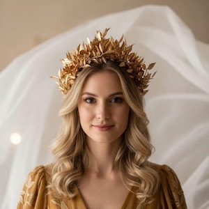 May include: A golden leaf crown, a headpiece, adorns a person's head. The crown is made of many small, detailed leaves. The person is wearing a mustard-colored dress with embroidered details. The background is a soft, neutral color.