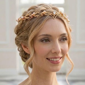 Può includere: Un copricapo in metallo color oro con un intricato design a filo intrecciato, impreziosito da piccole perle bianche rotonde e un singolo cristallo trasparente taglio marquise al centro. L'accessorio poggia sulla fronte e sulla testa.