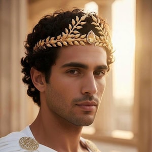 May include: A gold-colored laurel wreath crown with a central gemstone, worn by a person with dark curly hair. The crown is made of detailed leaf designs. The person is wearing a white garment with a gold circular brooch.