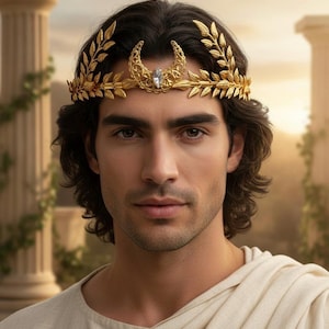 May include: A gold-colored laurel wreath crown with a crescent moon design and a central gemstone. The crown is worn by a person with dark hair and a cream-colored garment, set against a blurred background of columns and sunlight.