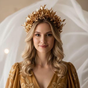 May include: A woman wearing a gold leaf crown and a mustard-colored dress with leaf embroidery. The crown is made of gold-colored leaves and sits atop wavy blonde hair. The dress has a V-neck and long sleeves.