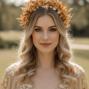 May include: A woman with blonde wavy hair wearing a gold leaf crown and a beige dress with gold leaf embroidery. The crown is made of multiple gold leaves. The background is a blurred green field.
