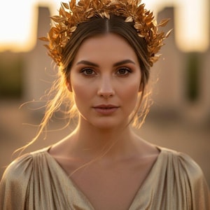 May include: A woman wearing a gold-colored leaf crown and a matching gold dress. The crown is made of multiple leaves. The dress has a V-neck and draped sleeves. The background is blurred with warm tones.