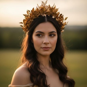 May include: A woman wearing a gold-colored leaf crown. The crown is made of multiple gold leaves. She is wearing a gold-colored off-the-shoulder dress. The background is a blurred green field and a soft, golden sky.