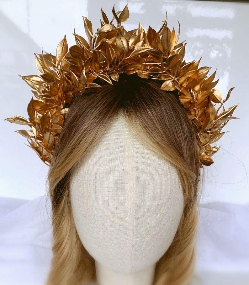 May include: A golden leaf crown, likely for a special occasion, sits atop a mannequin head with blonde hair. The crown is made of many gold-colored leaves, creating a full, textured appearance. The leaves are arranged in a circular shape, suggesting a headpiece.