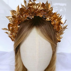 May include: A golden leaf crown, likely for a special occasion, sits atop a mannequin head with blonde hair. The crown is made of many gold-colored leaves, creating a full, textured appearance. The leaves are arranged in a circular shape.