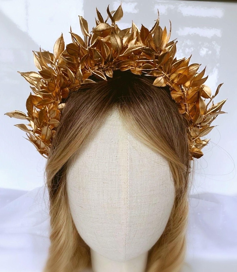 May include: A golden leaf crown, likely for a special occasion, sits atop a mannequin head with blonde hair. The crown is made of many gold-colored leaves, creating a full, textured appearance. The leaves are arranged in a circular shape.